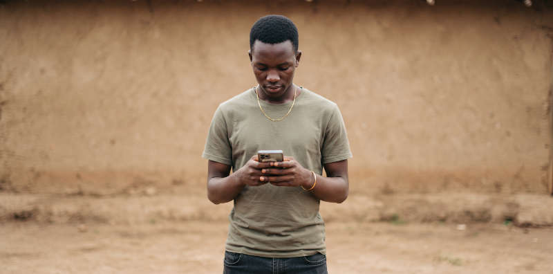 A young man standing at a mud wall, looking down at the phone in his hands