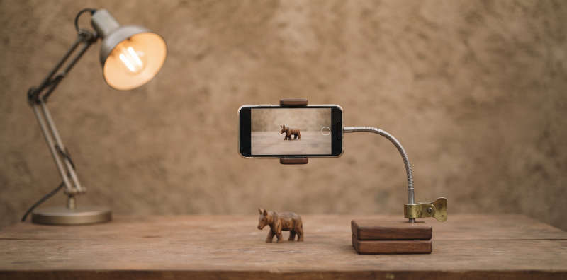 A phone on a homemade stand pointed at a carved wooden rhino on a table, a desk lamp to one side