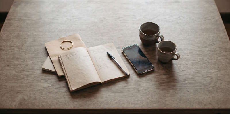 An open notebook with handwritten notes, a pen, a phone and two cups seen from directly above
