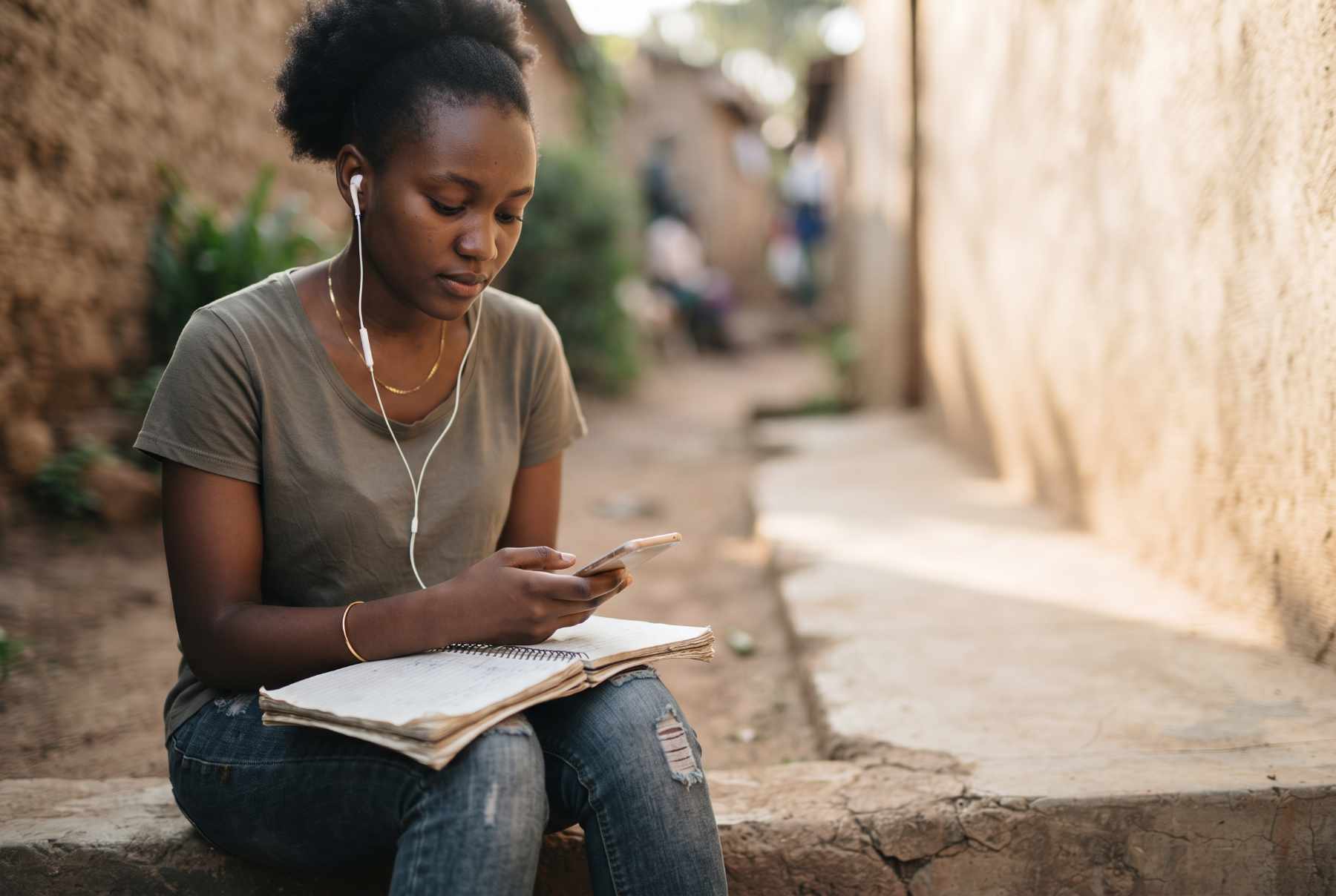 A young woman sitting on a step with earphones in, a phone in one hand and an exercise book on her lap